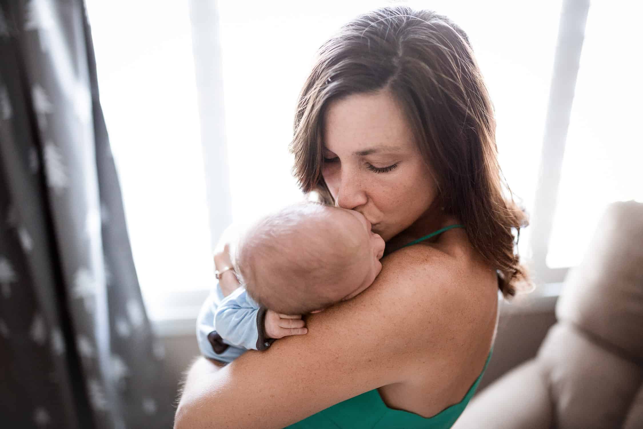 kansas city newborn photographer captures mom kissing her baby boy on his head in front of a window of the nursery